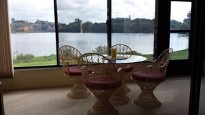 Quarto com cadeiras de vime e uma mesa, janelas amplas com vista para um lago e edifícios.