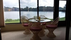 Una habitación con sillas de mimbre y una mesa, ventanas amplias con vistas a un lago y edificios.