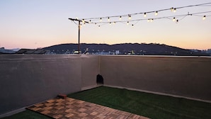 A rooftop terrace with a view of a cityscape at dusk, featuring a string of lights and a small patch of artificial grass. - Designer House (수원)