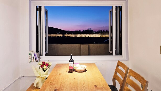 A dining table with a bottle of wine, a plate, and a bouquet of flowers in front of a window overlooking a cityscape at dusk. - Designer House (수원)