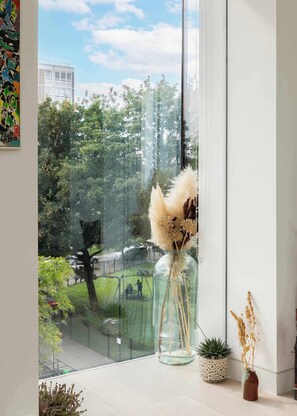 A window with a view of a park and a building, a vase with dried plants, and a small potted plant on the windowsill.