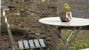 A metal-framed table with a potted plant on it, a white chair, and a brick wall in the background. - Sun-Drenched Shoreditch 2BR Penthouse with Balcony (London)