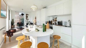 A modern kitchen with white cabinetry, a round island with stools, and a dining area with a sofa and coffee table. - Sun-Drenched Shoreditch 2BR Penthouse with Balcony (London)