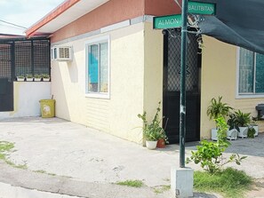 A residential building with a beige exterior, a black door, and a window with a blue curtain. There are potted plants on the ground and a street sign indicating 'Ballbtitan' and 'Almon' streets. - Amigas Transient House (Santo Tomas)