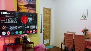 A living room with a flat-screen TV displaying a Welcome to Derry show, a large purple teddy bear, a wooden door, and a dining area with chairs and a table. - Amigas Transient House (Santo Tomas)