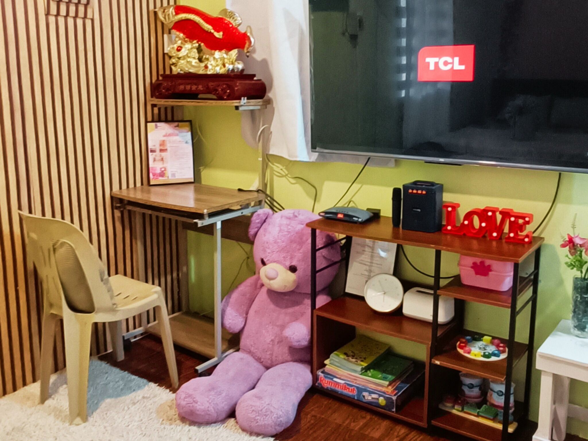 A room with a desk, a chair, a large teddy bear, a shelf with books and a clock, and a TCL TV.