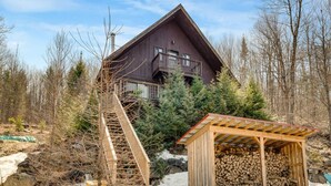 A wooden cabin with a balcony, a wooden staircase, and a covered area for storing firewood. - Woodsy Chalet Near Tremblant (Amherst)