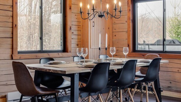A dining area with a wooden table set for four, surrounded by black chairs. A chandelier hangs above the table, and there are two windows providing a view of the outside. - Woodsy Chalet Near Tremblant (Amherst)