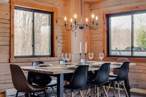 A dining area with a wooden table set for four, surrounded by black chairs. A chandelier hangs above the table, and there are two windows providing a view of the outside.