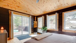 A cozy bedroom with a large bed, wooden walls, and a view of a snowy landscape through the windows. - Woodsy Chalet Near Tremblant (Amherst)
