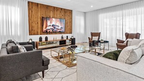 A modern living room with a large TV mounted on a wooden wall, a glass coffee table, and comfortable seating. - 9BR house sleeps 21 with spa and pool table (Kissimmee)