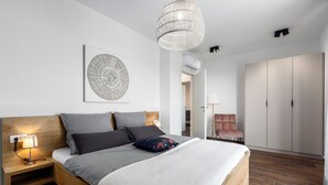 Une chambre moderne avec une tête de lit en bois, un grand lit avec une literie grise, une œuvre d’art abstraite accrochée au mur, un luminaire suspendu et une armoire.