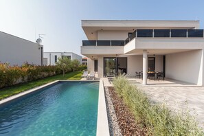 Una casa moderna de dos pisos con piscina, mobiliario exterior y un jardín cuidado.
