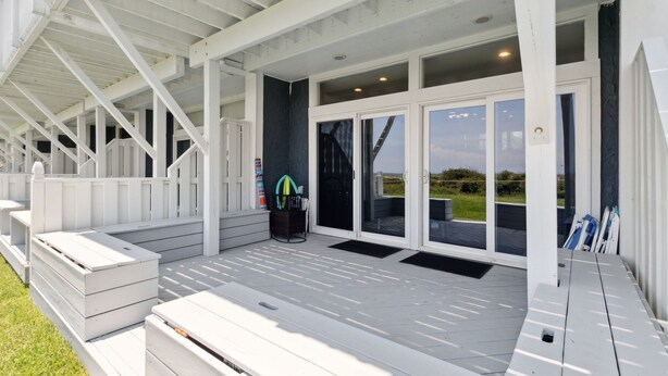 A covered outdoor area with a wooden deck, a black trash bin, and a set of outdoor furniture. - Sandpiper Sunrise - Hosted by Moore AI Rentals (Fernandina Beach)