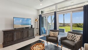 A spacious living room with a large TV, comfortable seating, and a view of the beach through large windows. - Sandpiper Sunrise - Hosted by Moore AI Rentals (Fernandina Beach)