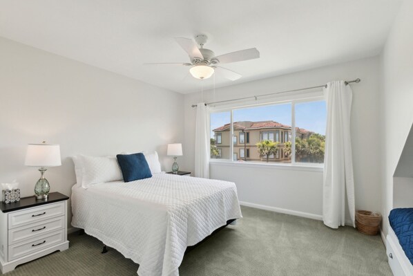 A bedroom with a bed, nightstands, a ceiling fan, a window with curtains, and a view of buildings outside. - Sandpiper Sunrise - Hosted by Moore AI Rentals (Fernandina Beach)