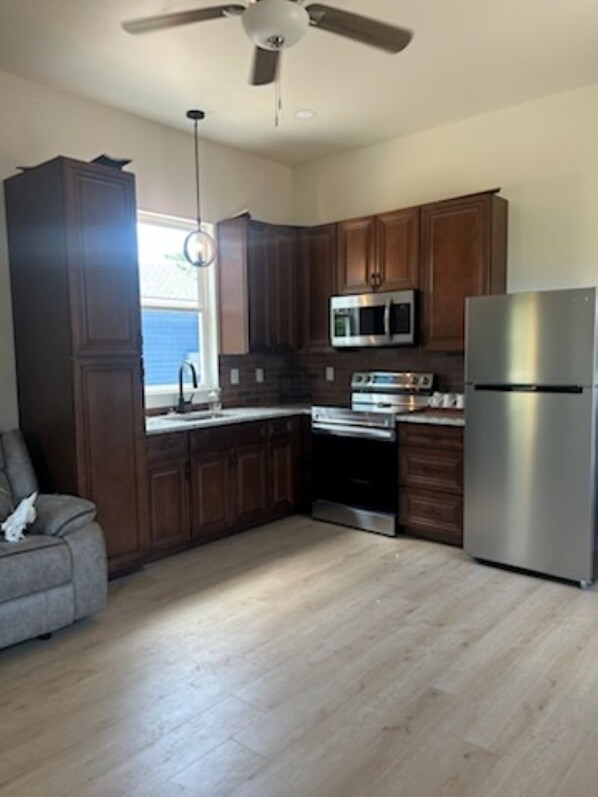 A kitchen with dark wooden cabinets, a stainless steel refrigerator, a microwave, and a stove. There is a ceiling fan and a window with a view of the sky.