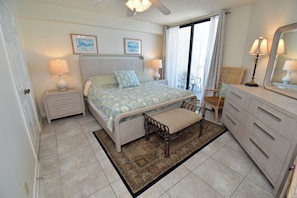 Un dormitorio con cama, mesitas de noche, cómoda, banco, ventilador de techo y vistas al exterior.
