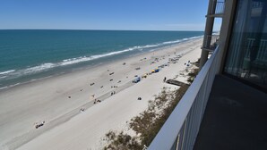 On peut admirer la plage depuis un balcon donnant sur une étendue de sable, avec des parasols et des gens.