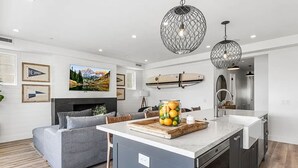A modern kitchen with a central island, a grey sofa, and a dining area. There are framed pictures on the walls and a surfboard decoration. - 3BR Loft Ocean View Rooftop Hot Tub Parking (Newport Beach)