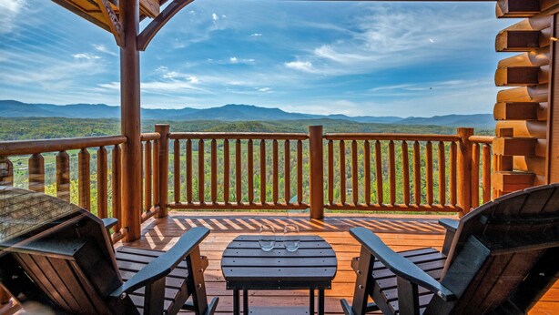 A wooden deck with a view of mountains, two chairs, and a table with glasses on it. - Breathtaking Mountain Views • Resort Pool • Hot Tub • Sauna • Near Pigeon Forge (Sevierville)
