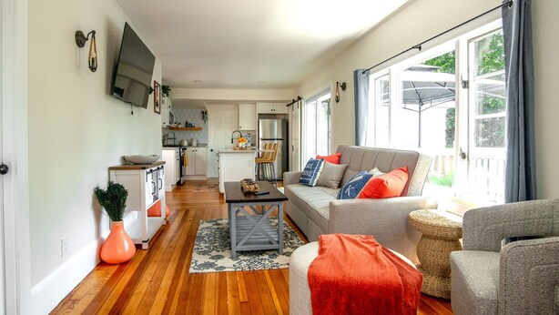 A spacious living room with a grey sofa, a coffee table, and a red throw blanket. There is a kitchen in the background with white cabinets and a flat-screen TV mounted on the wall. - Lakeview Getaway With Hot Tub (Billerica)
