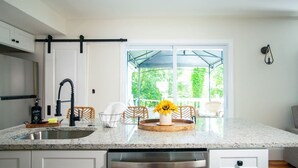 A modern kitchen with a stainless steel dishwasher, white cabinets, and a large window with a view of greenery. - Lakeview Getaway With Hot Tub (Billerica)