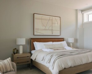 A neatly arranged bedroom with a large bed, bedside lamps, a nightstand, a chair, and a wall-mounted abstract artwork.