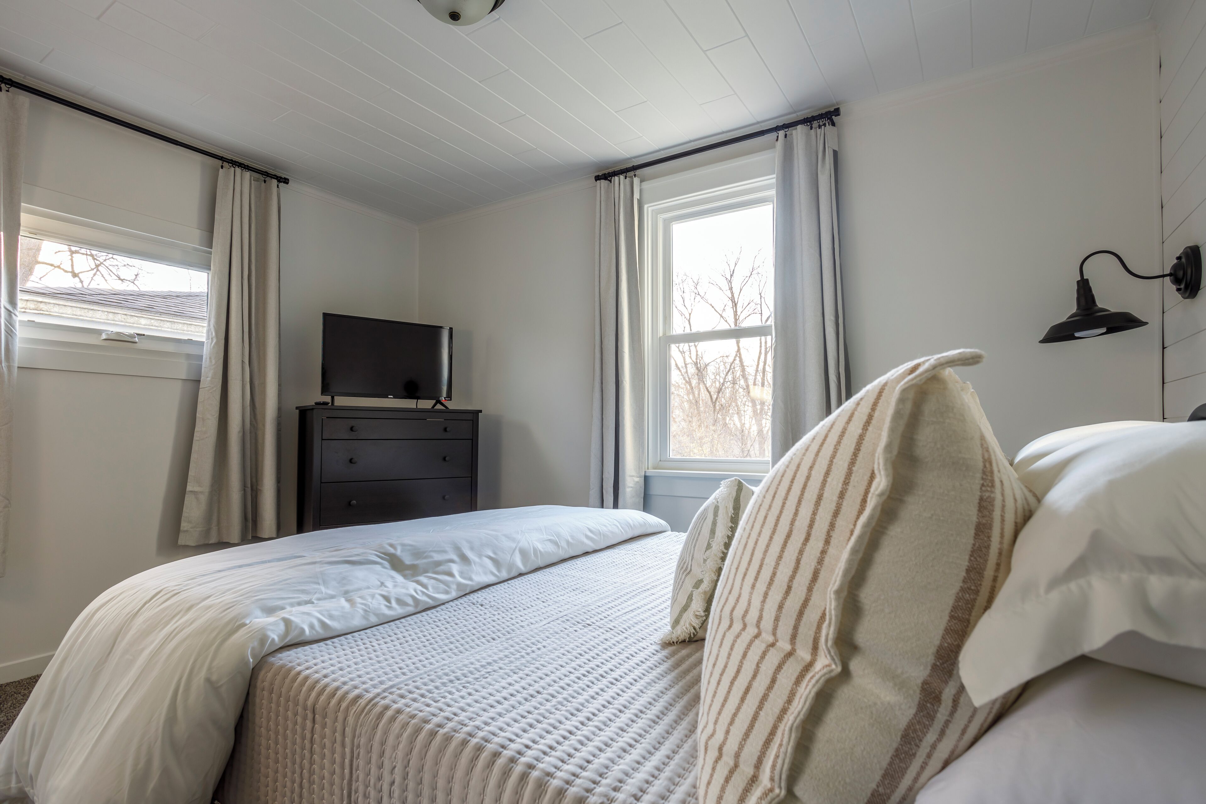 Un dormitorio con cama, cómoda, televisor y ventana con cortinas.