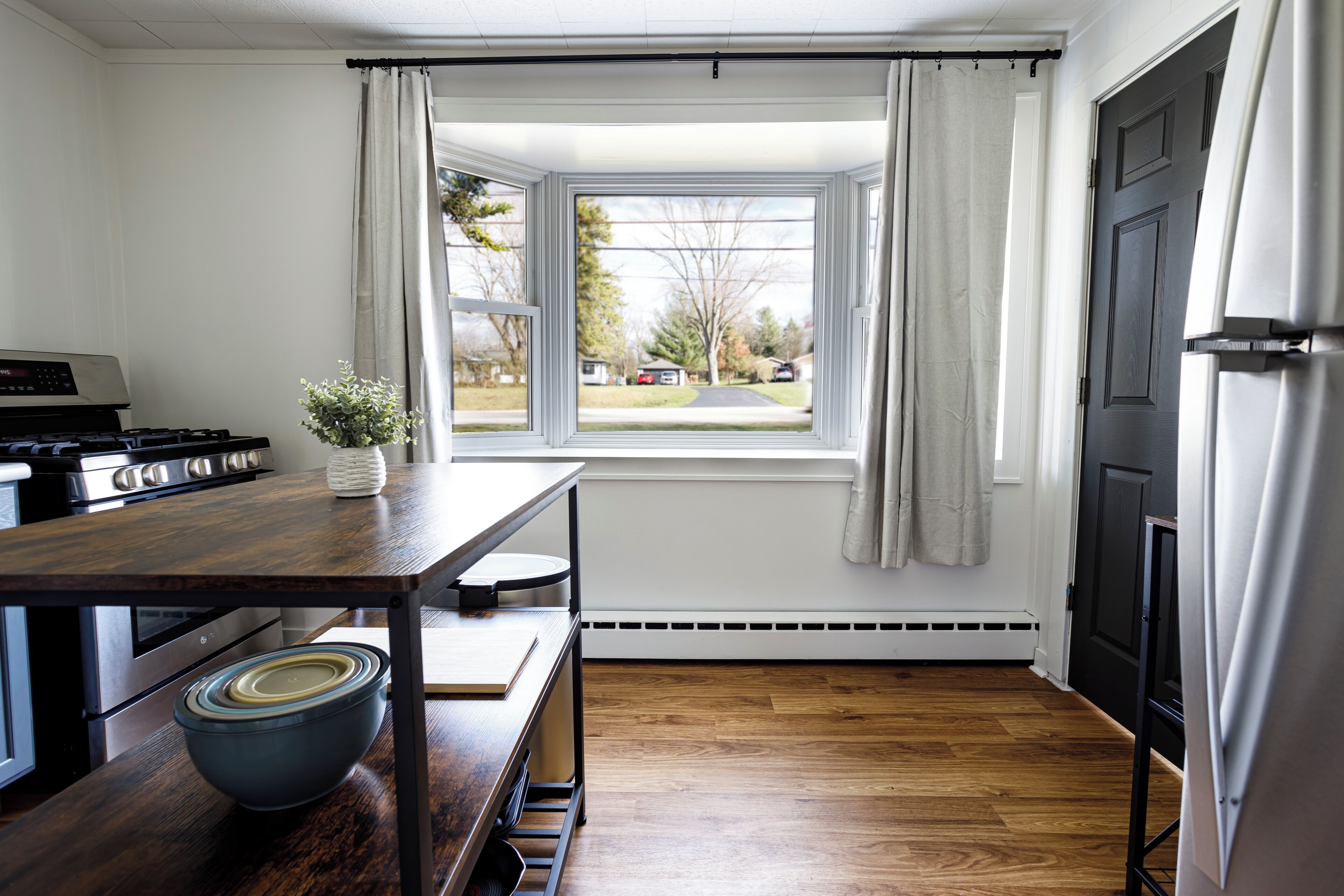 Een keuken met een houten tafel, een kom, een fornuis en een raam met gordijnen.