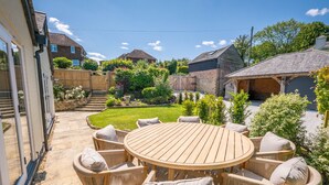 Une terrasse avec une table ronde en bois et des chaises en osier, entourée d’un jardin bien entretenu et, au fond, d’une maison en briques.