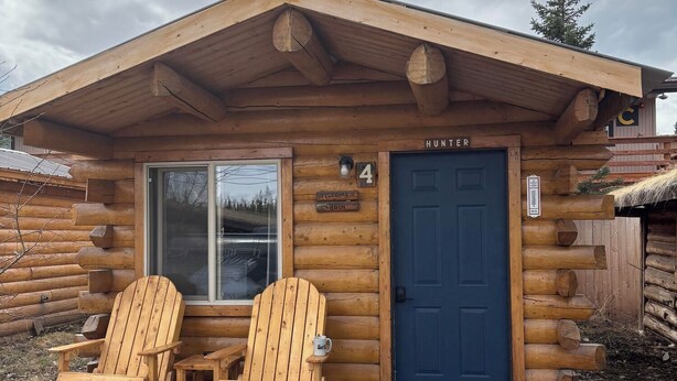 Eine Blockhütte mit zwei Holz-Adirondack-Stühlen und einem kleinen Tisch vor der Tür, einer blauen Tür und einem Schild mit der Aufschrift 'Willkommen in Hütte 4'.