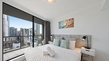 A modern hotel room with a large window, a bed with white linens, a gray headboard, a nightstand with a lamp, and a cityscape view.