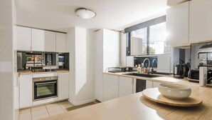 A modern kitchen with white cabinetry, a built-in oven, a sink, and a large window. - KozyGuru | Homebush West | Modern 2R Apt + Parking (Homebush West)