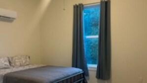 Une chambre à coucher avec un lit, un ventilateur de plafond, une fenêtre avec des rideaux et une vue sur les arbres à l’extérieur.