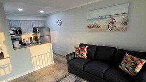 A living room with a black sofa, two patterned cushions, a rug, and a painting of a bicycle on a beach. - Sunset paradise, entire rental unit in Tampa,florida (Tampa)