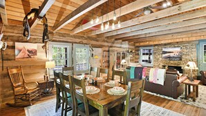 Interior rústico de una cabaña con comedor, muebles de madera, chimenea de piedra y ventilador de techo.