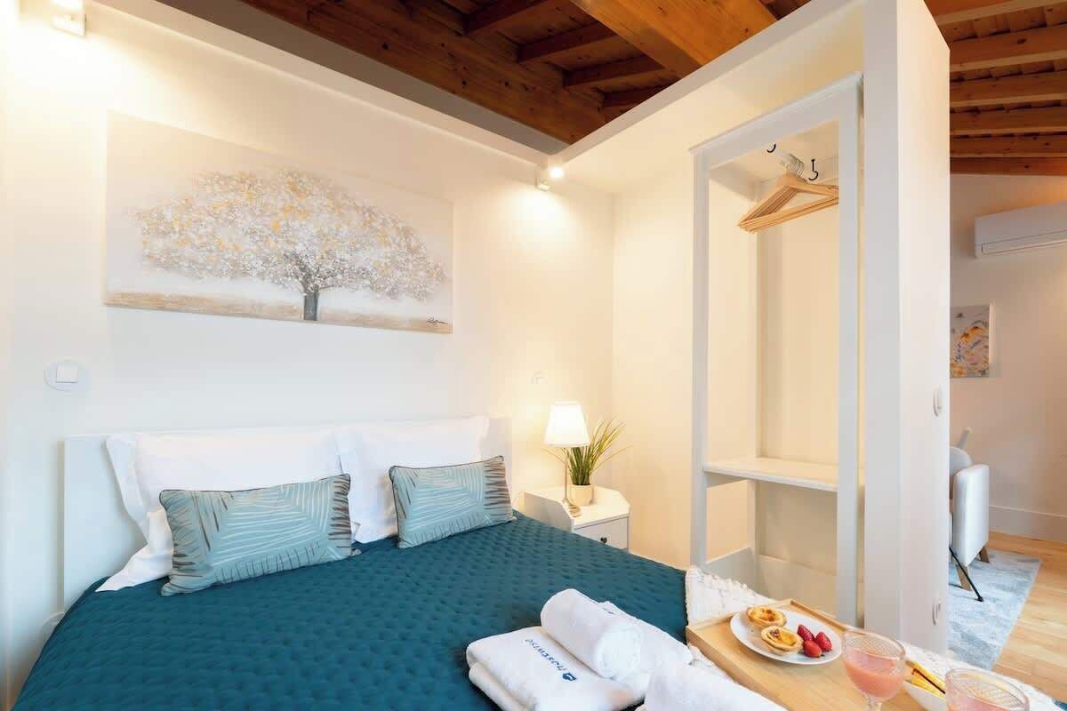 A bedroom with a bed, pillows, a bedside table with a lamp, a painting on the wall, and a tray with food and drinks.