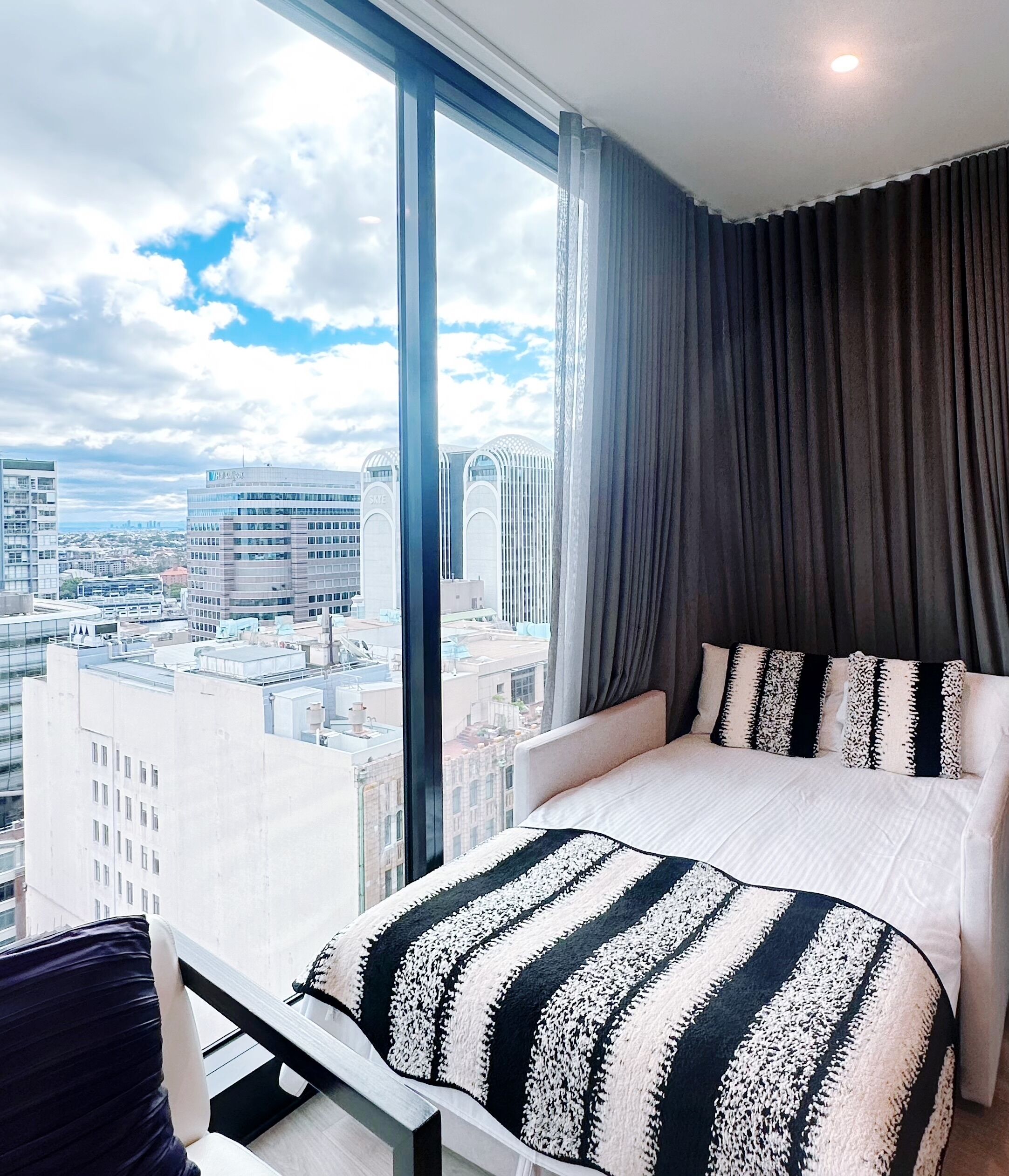 Habitación de hotel moderna con un ventanal que ofrece vistas a la ciudad, una cama bien hecha con ropa de cama a rayas y una cortina oscura que cubre la ventana.