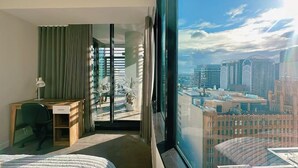 A hotel room with a bed, desk, and chair, a view of the cityscape through large windows. - Stylish 1BR suite with City-view & Balcony (Sydney)