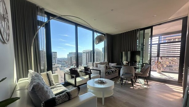 A modern living room with a sofa, coffee table, and chairs, large windows with a city view, and a flat-screen TV. - Stylish 1BR suite with City-view & Balcony (Sydney)