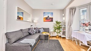 Una sala de estar moderna con un sofá gris, una mesa redonda pequeña, una planta en maceta y una zona de comedor con una mesa para dos personas.