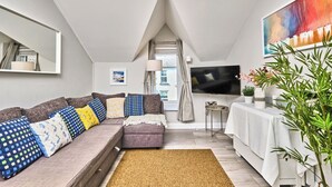 A modern living room with a sectional sofa, a flat-screen TV, a small table, and a large window. - Central Portrush Penthouse Retreat (Portrush)