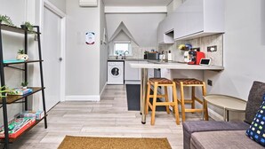 A compact living space with a kitchenette, a washing machine, a dining area with stools, and a small seating area with a sofa and a coffee machine. - Central Portrush Penthouse Retreat (Portrush)