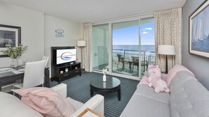 A hotel room with a balcony, a television, a dining area, and a sofa with a plush toy.