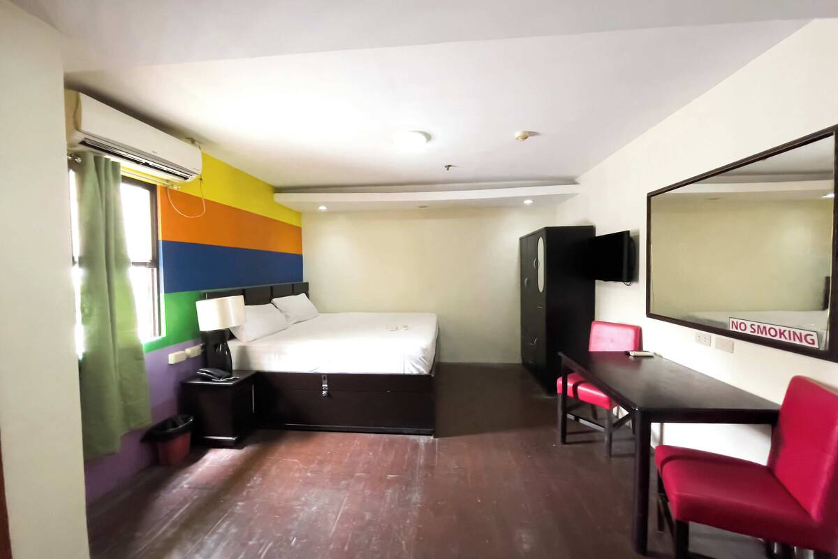 Quarto de hotel com cama, escrivaninha, cadeira, televisão e guarda-roupa.