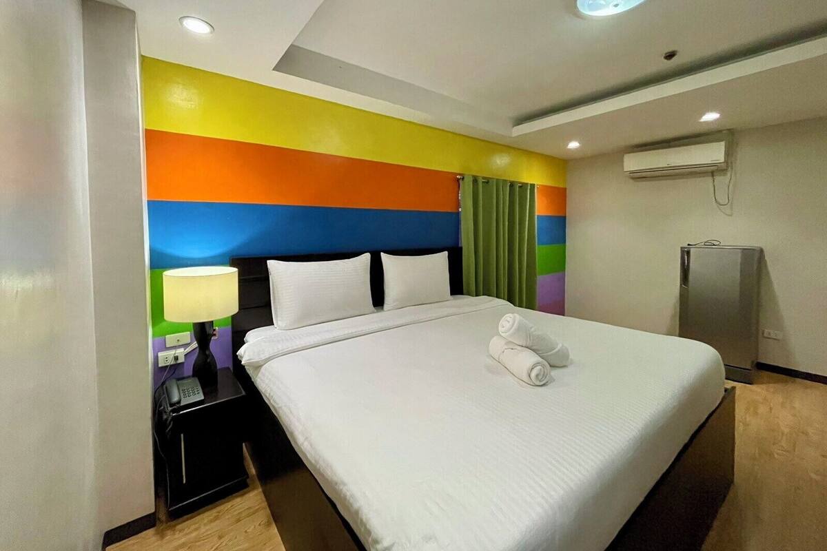 Quarto de hotel com cama, mesa de cabeceira com luminária, telefone e geladeira.