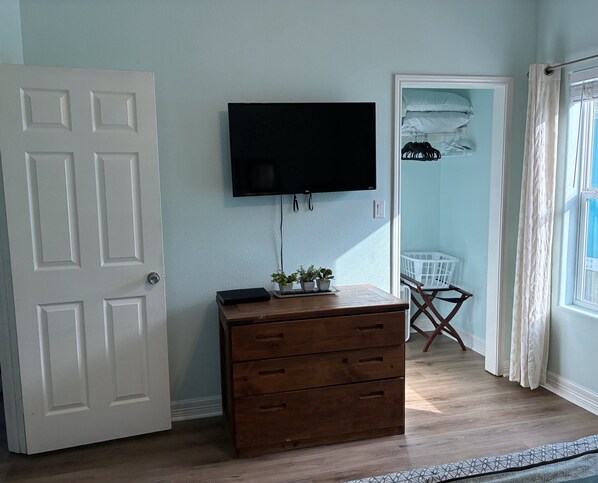Una habitación con un aparador de madera, un televisor de pantalla plana montado en la pared, una puerta, una ventana con cortinas y un cesto de ropa en la esquina.