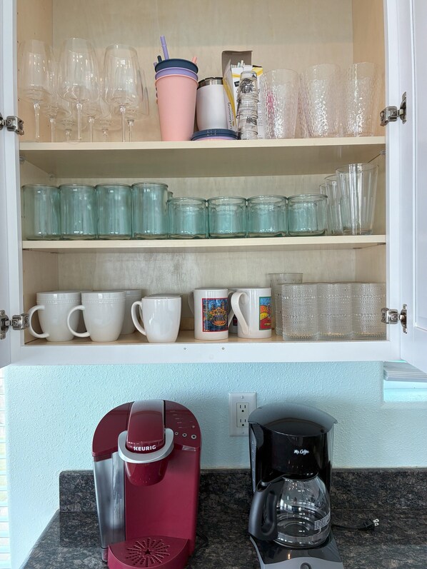 Una cocina con un armario que contiene varios vasos y tazas, una cafetera Keurig roja y una cafetera Mr. Coffee negra.