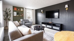 A modern living room with a grey sofa, a round coffee table, a TV mounted on the wall, and a bookshelf. - King Size Bed, Work Desk, Near Hwy, Laundry Room Access, Nearby Parking (Lockport)